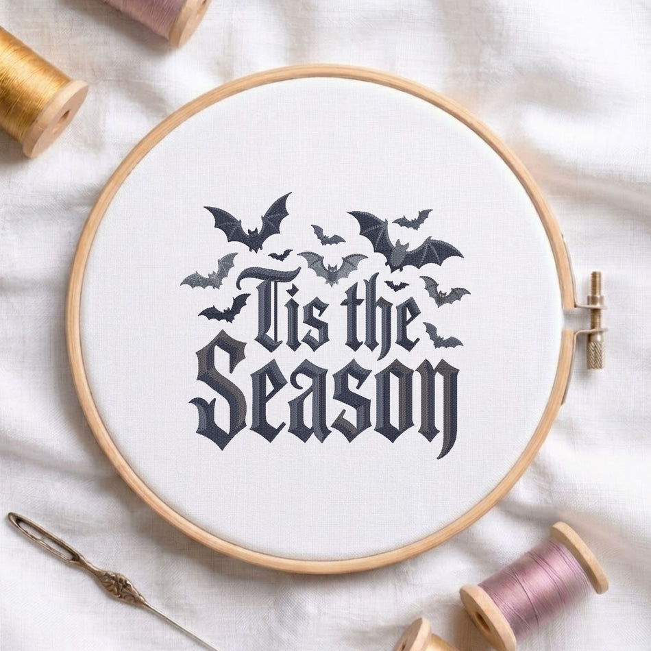 A circular embroidery hoop holding white fabric with a gothic-style black design that reads 'This is the Season' beneath several flying bats. The hoop is styled on a white fabric surface with spools of thread and a needle around it.