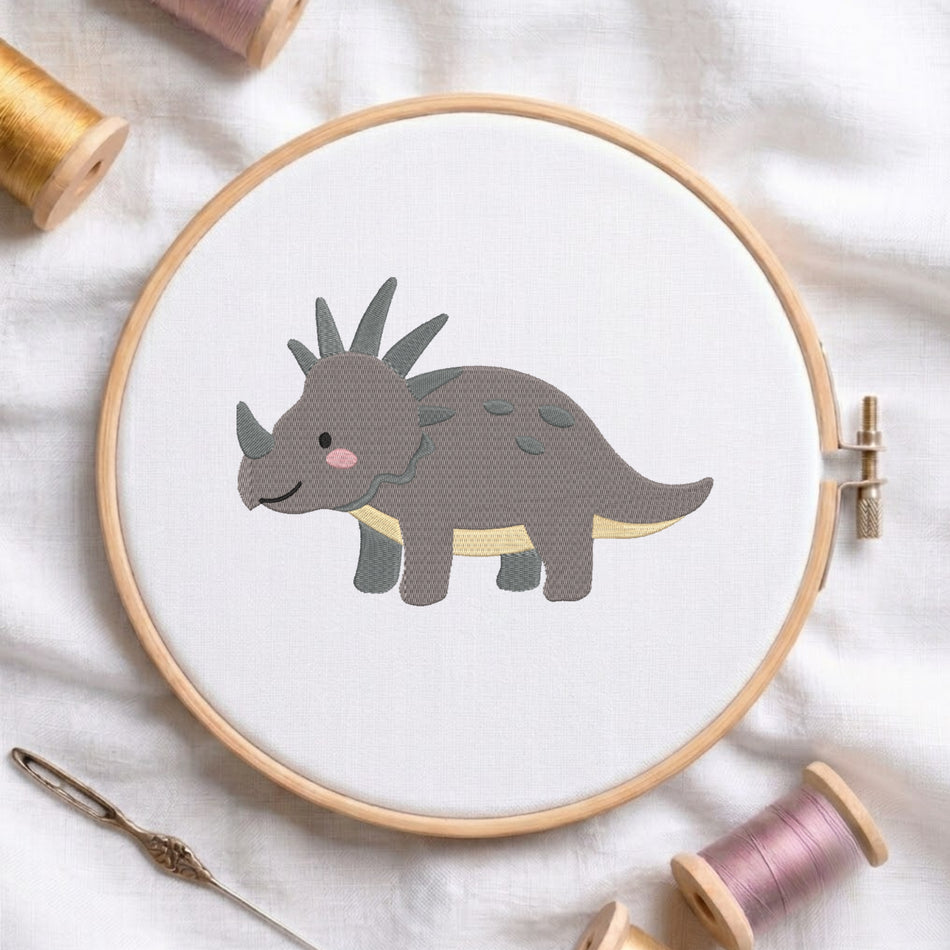 A wooden embroidery hoop holds white fabric with a stitched cartoon Triceratops dinosaur in gray, black, pink, and pale yellow. The hoop is styled on a white fabric surface with spools of thread and a needle around it.