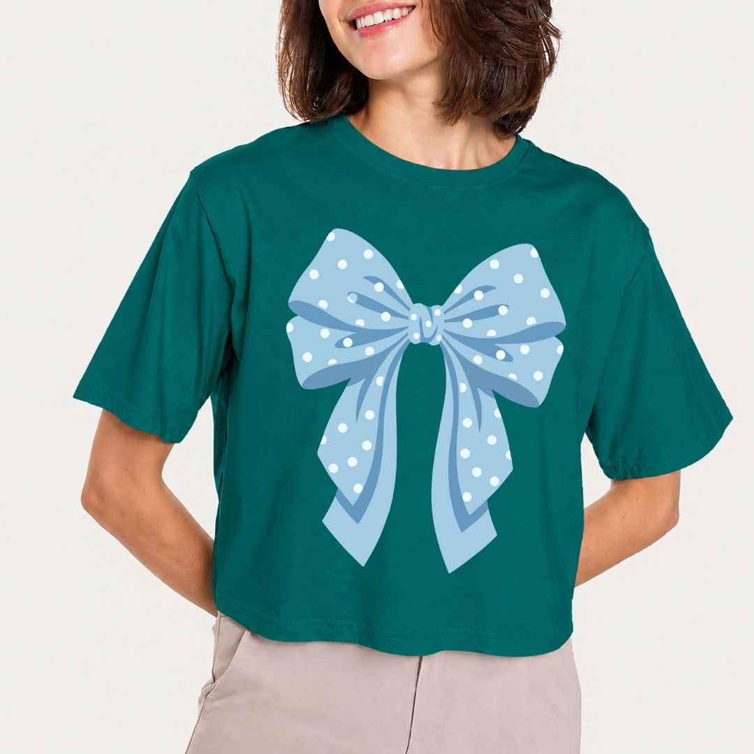 A woman wearing a dark teal cropped t-shirt with a large light blue polka dot bow graphic on the front, paired with beige pants, against a plain off-white background.