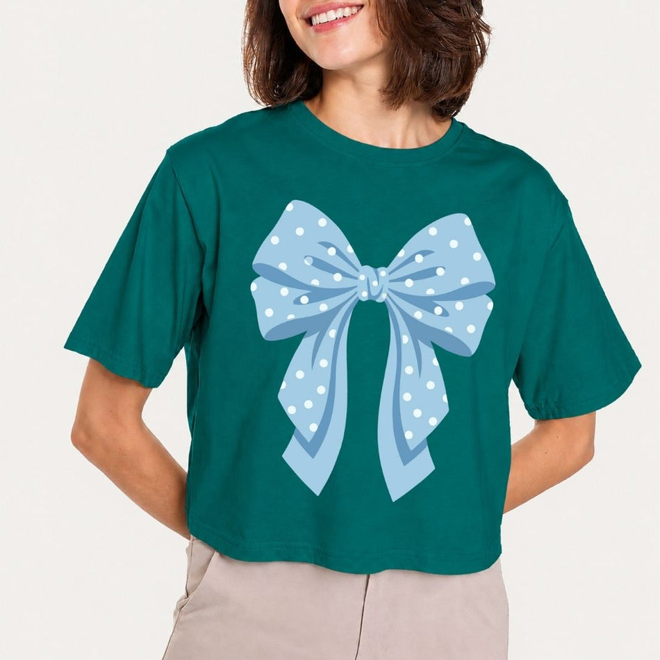 A woman wearing a dark teal cropped t-shirt with a large light blue polka dot bow graphic on the front, paired with beige pants, against a plain off-white background.