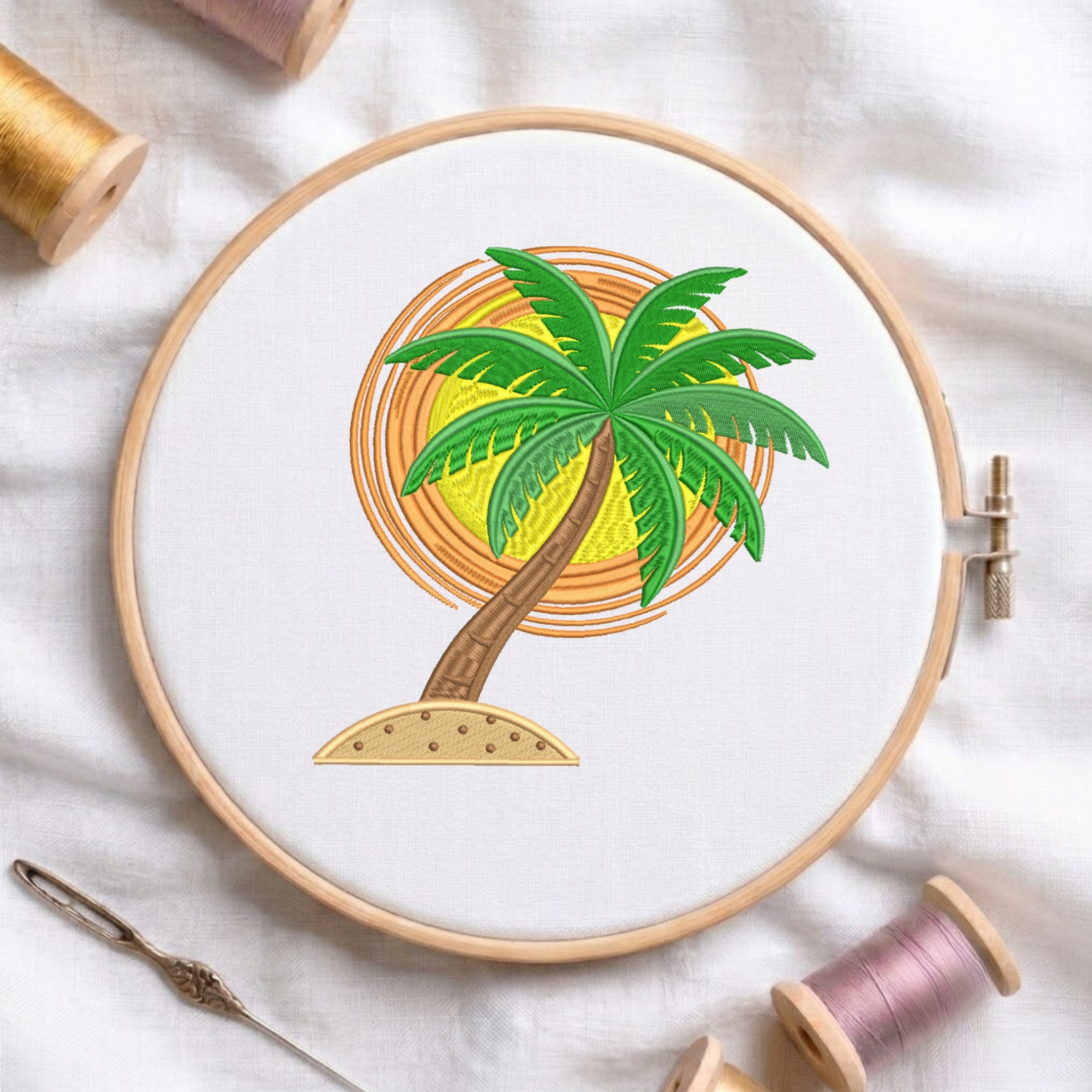 A wooden embroidery hoop holds white fabric with a palm tree design in front of an orange and yellow sun motif. The hoop is styled on a white fabric background with spools of thread and a needle nearby.