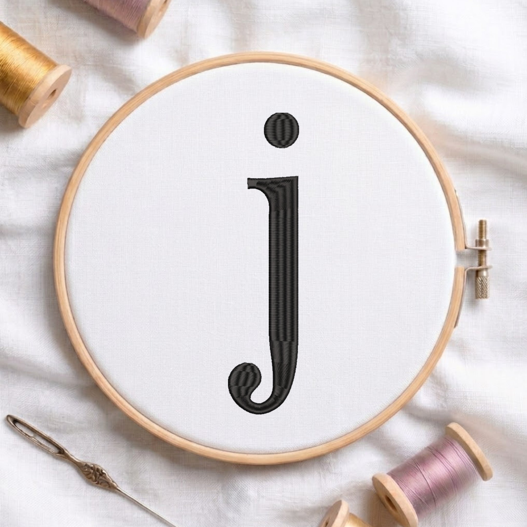 A wooden embroidery hoop holding white fabric with a large black embroidered letter 'j', surrounded by spools of thread and a needle on a white fabric background.