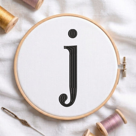 A wooden embroidery hoop holding white fabric with a large black embroidered letter 'j', surrounded by spools of thread and a needle on a white fabric background.