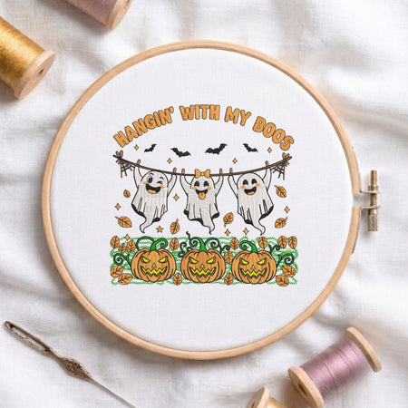 A circular embroidery hoop holding white fabric with a Halloween-themed stitched design. The design shows three smiling ghosts hanging from a line with pumpkins, bats, leaves, and the phrase “HANGIN’ WITH MY BOOS” in orange.