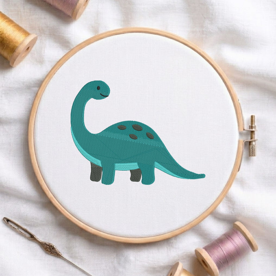 A wooden embroidery hoop holds white fabric with a teal dinosaur design stitched in the center. The hoop is styled on a white fabric background with spools of thread and a needle around it.
