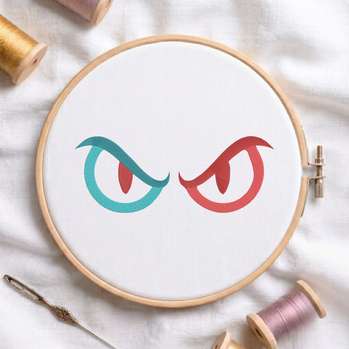 A circular embroidery hoop holding white fabric with a stylized pair of eyes design in teal and coral red. The hoop is photographed on a white fabric background with spools of thread and a needle around it.