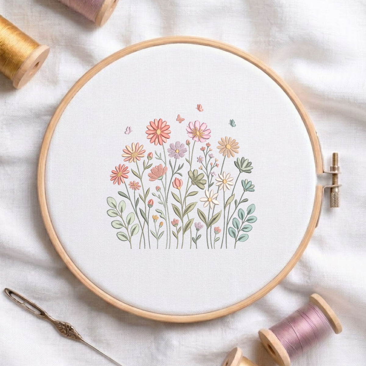 Cute pink, peach, and yellow wildflowers meadow with tiny butterflies machine embroidery design, displayed inside a round wooden hoop on white fabric with thread spools and a needle.
