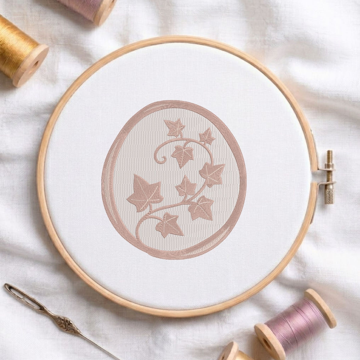 A round embroidery hoop holding white fabric with a dusty rose embroidered ivy design, surrounded by spools of thread and a needle on a white fabric background.