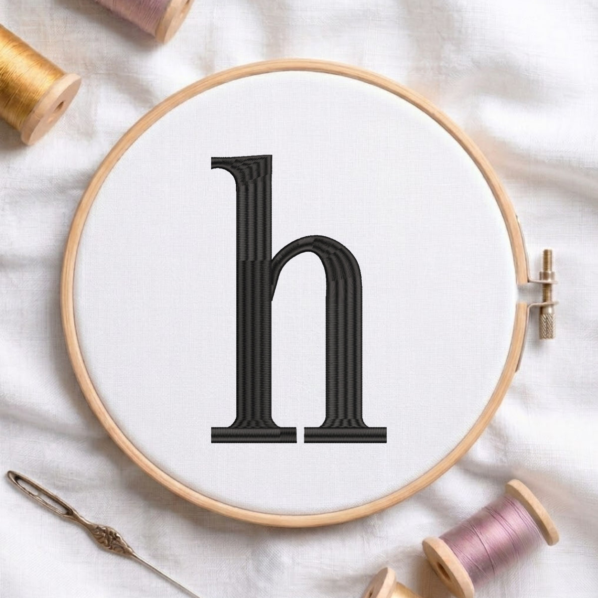 A wooden embroidery hoop holding white fabric with a black serif letter 'h' stitched in the center, surrounded by spools of thread and a needle on a white fabric background.