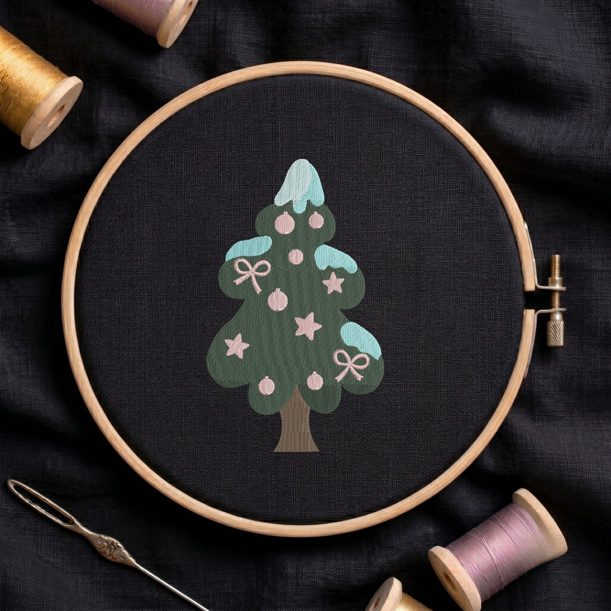 Christmas tree machine embroidery design featuring a green tiered body with snowy blue caps, decorated with pink stars, ornaments, and bows, displayed inside a round wooden hoop on black linen fabric, accompanied by wooden thread spools and a needle.