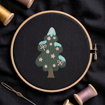 Christmas tree machine embroidery design featuring a green tiered body with snowy blue caps, decorated with pink stars, ornaments, and bows, displayed inside a round wooden hoop on black linen fabric, accompanied by wooden thread spools and a needle.
