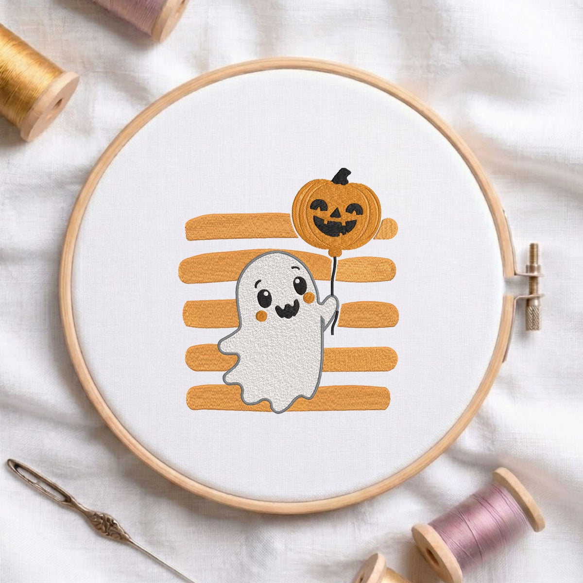 Cute ghost holding a pumpkin balloon machine embroidery design displayed in a wooden hoop on white fabric with thread spools and a needle