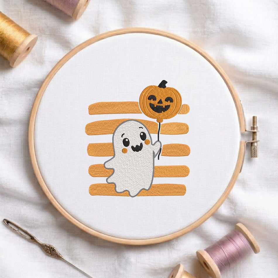 Cute ghost holding a pumpkin balloon machine embroidery design displayed in a wooden hoop on white fabric with thread spools and a needle