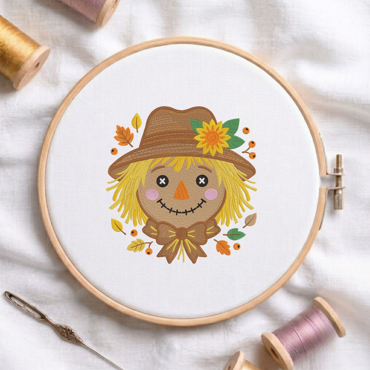 Cute autumn scarecrow face wearing a brown hat, sunflower, and fall leaves machine embroidery design shown in a wooden embroidery hoop on white fabric with thread spools and a needle