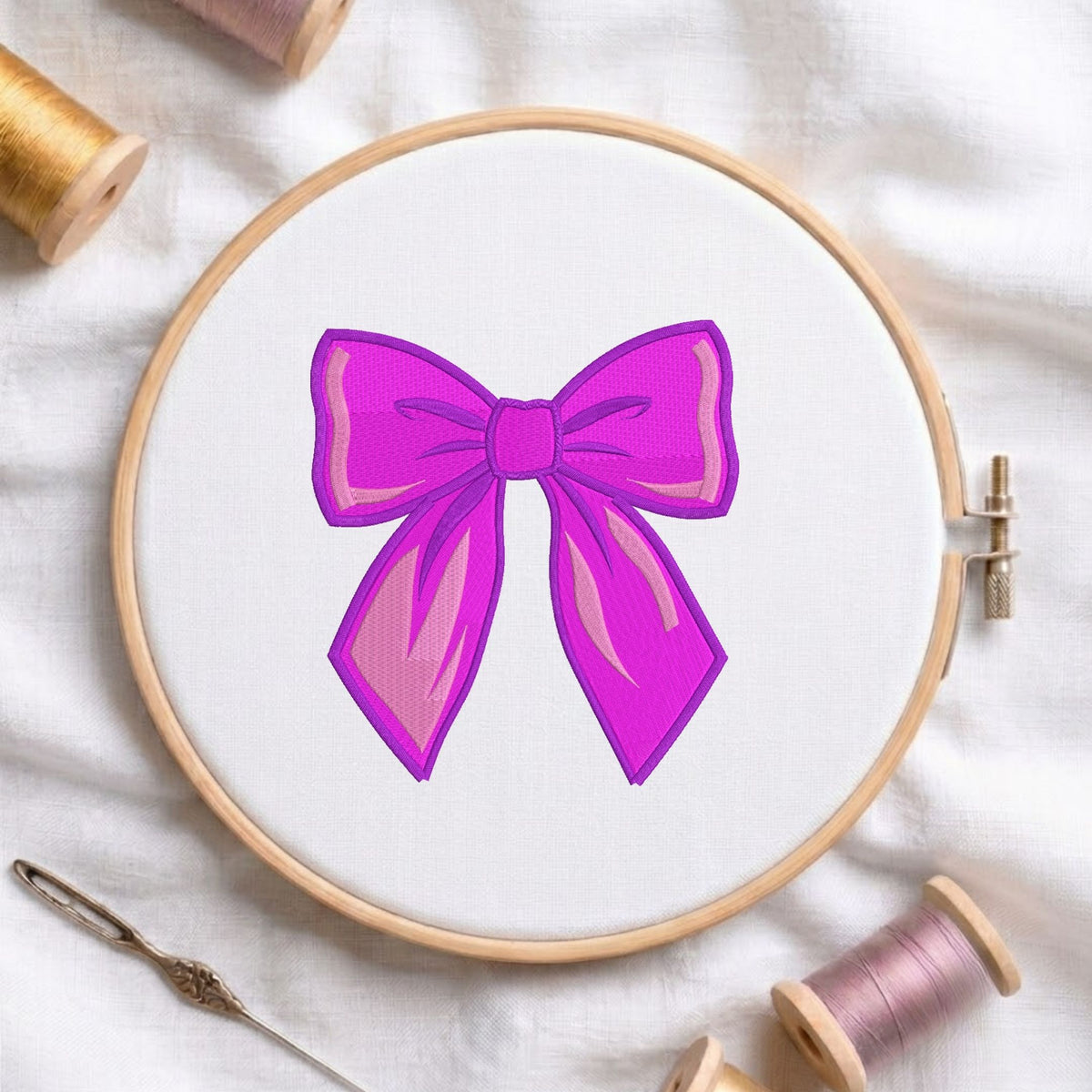 Vibrant purple bow machine embroidery design displayed inside a round wooden hoop on white fabric alongside thread spools and a needle.