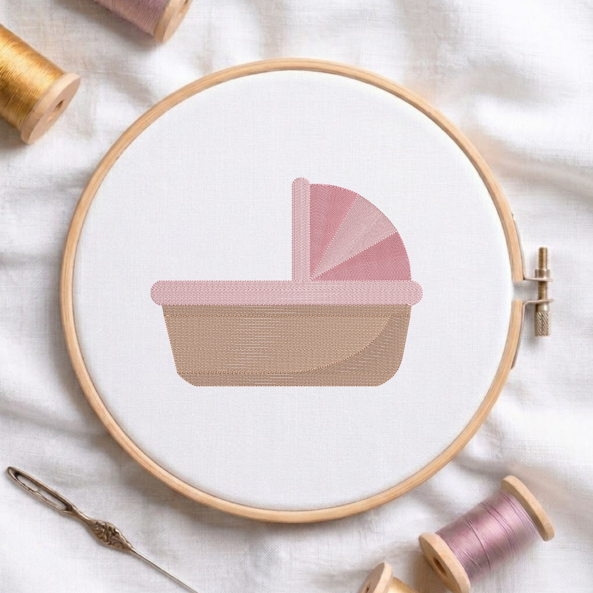 A circular embroidery hoop holding white fabric with a stitched baby carriage design in soft pink and beige. A needle and thread spools are arranged around it on a white fabric background.