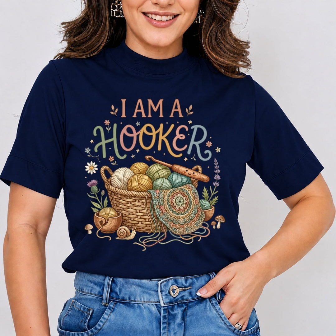 A woman wearing a navy blue graphic t-shirt with the text 'I AM A HOOKER' and a colorful illustration of a basket filled with yarn balls, crochet hooks, and knitting needles. She is also wearing blue jeans.