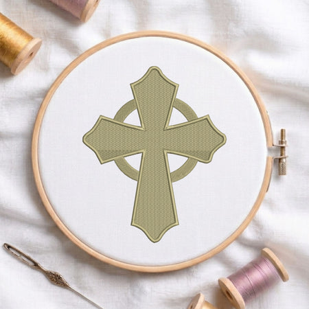 An embroidery hoop on a white cloth displays a finished machine embroidery design of a sage green Celtic cross with flared tips and an intersecting ring, flanked by spools of thread and a needle.