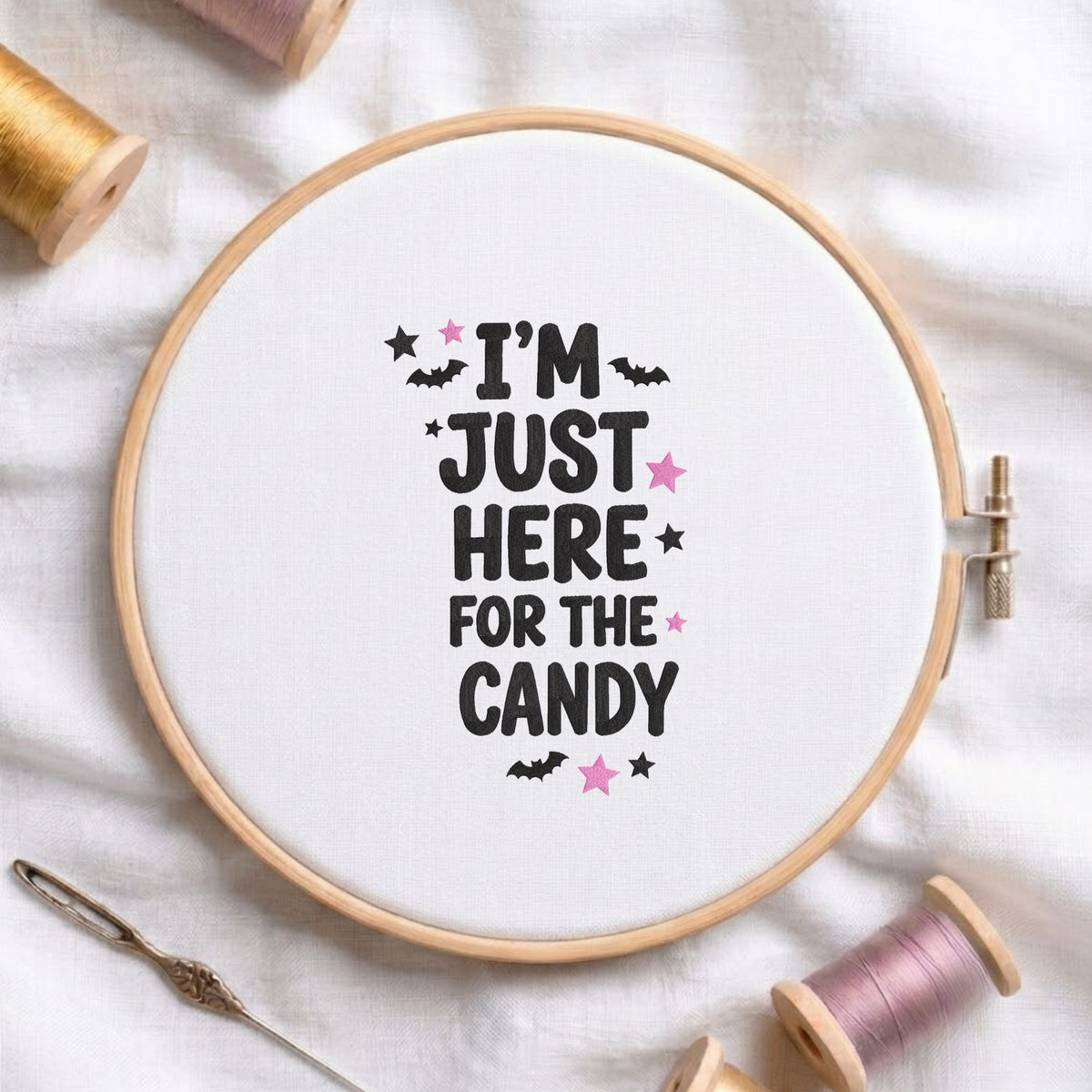 I'm Just Here for the Candy" Halloween quote with bats and stars machine embroidery design displayed in a wooden embroidery hoop on white fabric with thread spools and a needle