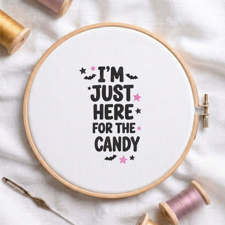 I'm Just Here for the Candy" Halloween quote with bats and stars machine embroidery design displayed in a wooden embroidery hoop on white fabric with thread spools and a needle