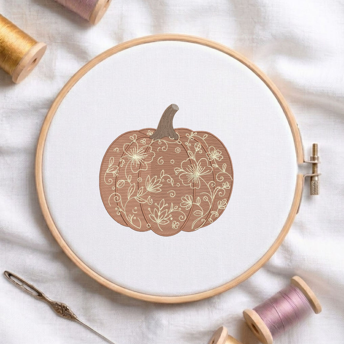 Floral vine-patterned pumpkin machine embroidery design displayed in a wooden hoop on white fabric with thread spools and a needle.