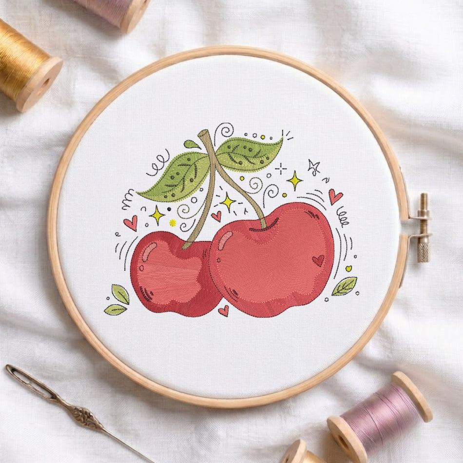 A circular embroidery hoop on white fabric, featuring a stitched design of two red cherries with green leaves and small decorative doodles. Spools of thread and a needle are arranged around the hoop.