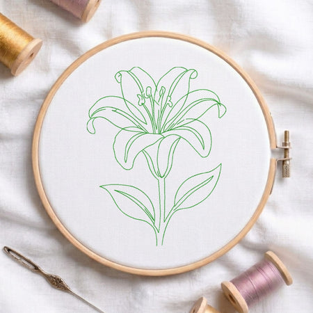 A wooden embroidery hoop holding white fabric with a green outline drawing of a lily flower. Spools of thread and a needle are arranged around it on a white fabric background.