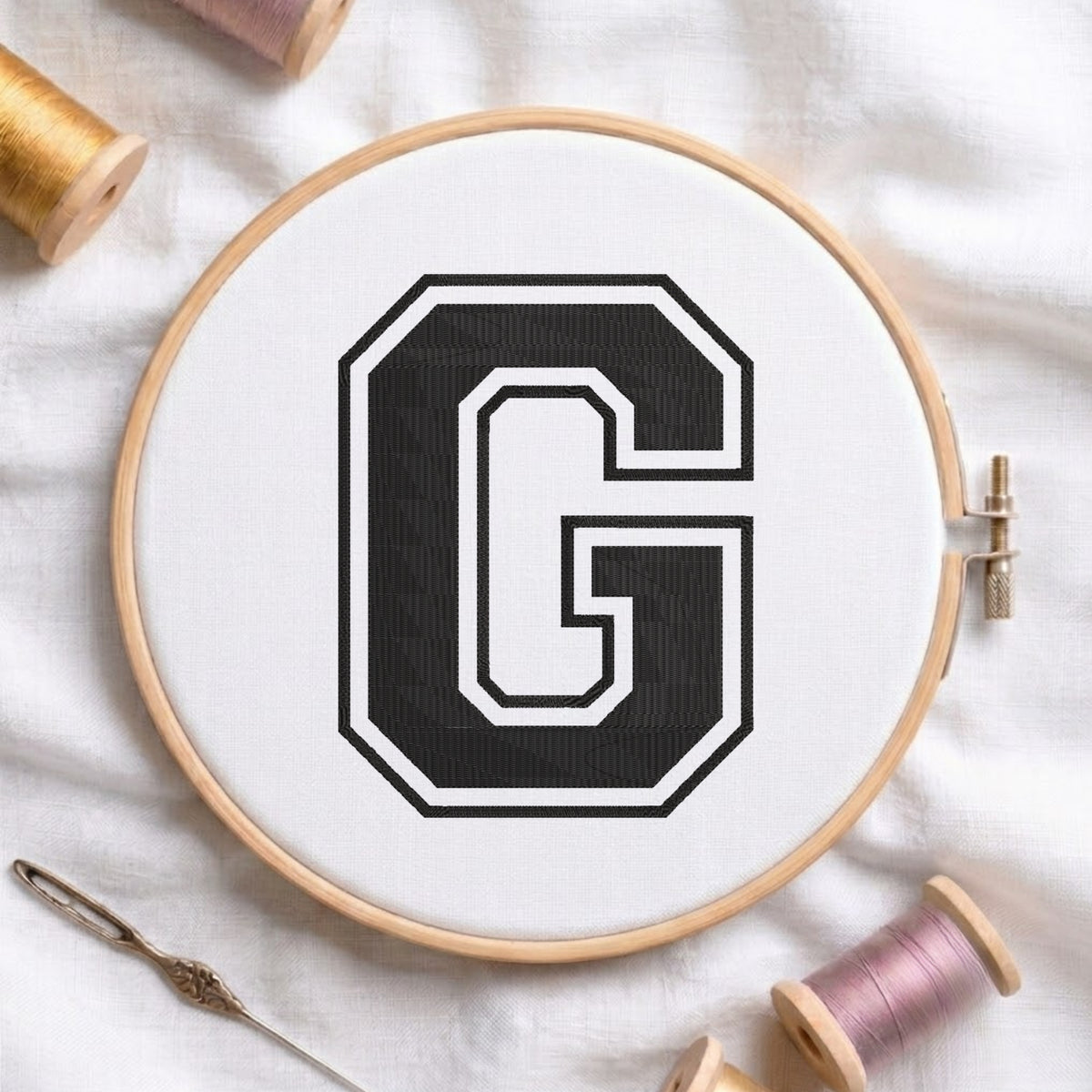 A wooden embroidery hoop holding white fabric with a large black letter G outlined in white, surrounded by spools of thread and a needle on a white fabric background.