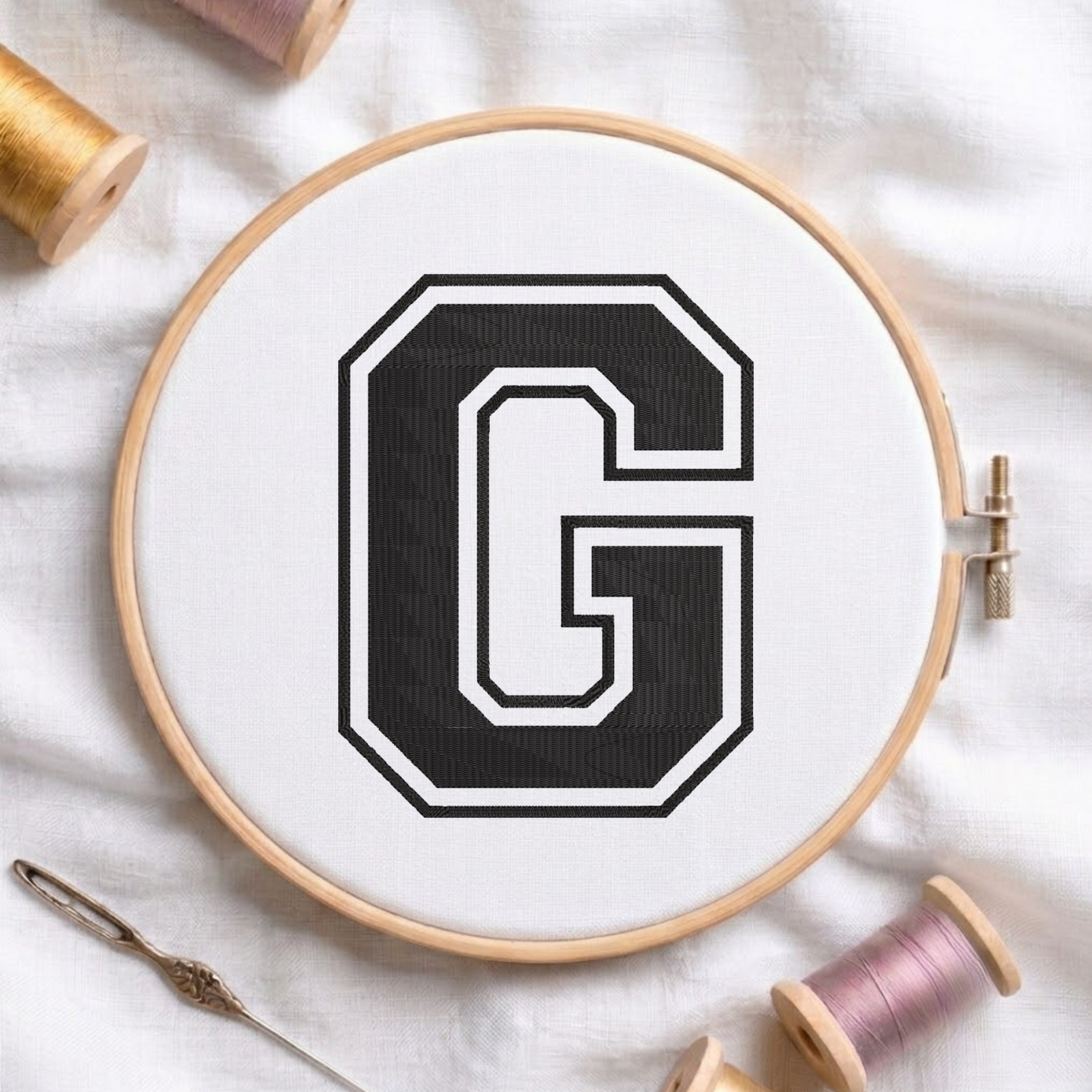 A wooden embroidery hoop holding white fabric with a large black letter G outlined in white, surrounded by spools of thread and a needle on a white fabric background.