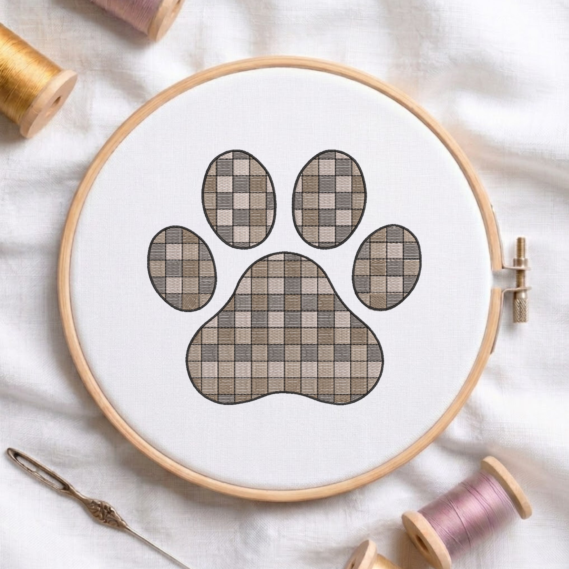 A circular embroidery hoop holding white fabric with a paw print design filled with a beige and taupe checkerboard pattern. Spools of thread and a needle are arranged around it on a white fabric background.