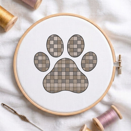 A circular embroidery hoop holding white fabric with a paw print design filled with a beige and taupe checkerboard pattern. Spools of thread and a needle are arranged around it on a white fabric background.