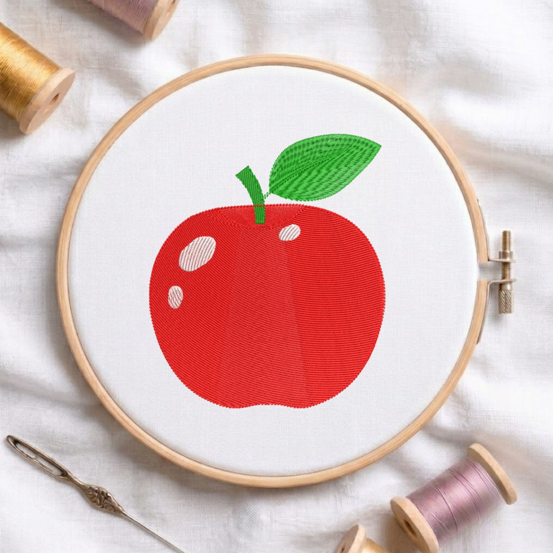 A wooden embroidery hoop holding white fabric with a stitched red apple design and green leaf, surrounded by spools of thread and a needle on a white fabric background.
