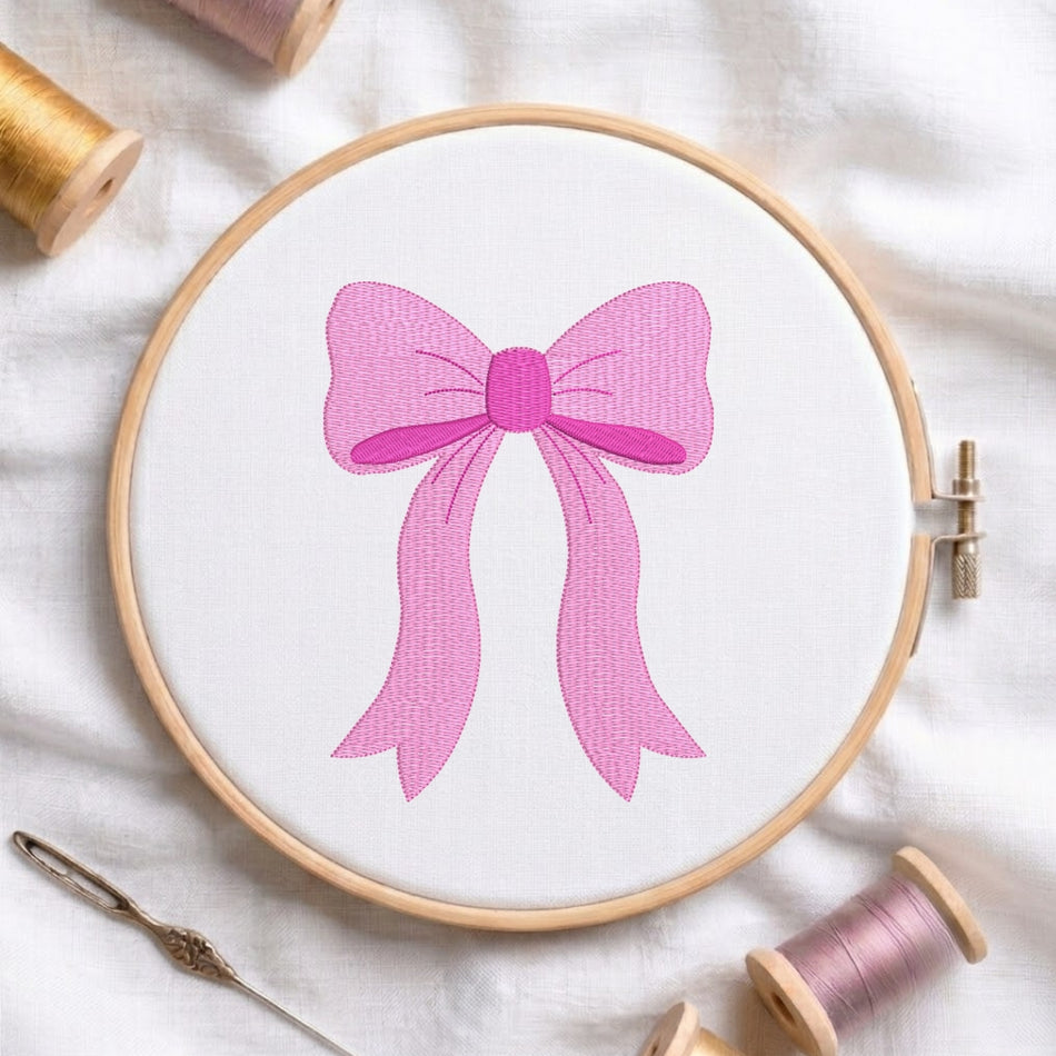 A pink bow embroidery design stitched on white fabric inside a wooden hoop, surrounded by spools of thread and a needle on a white fabric background.