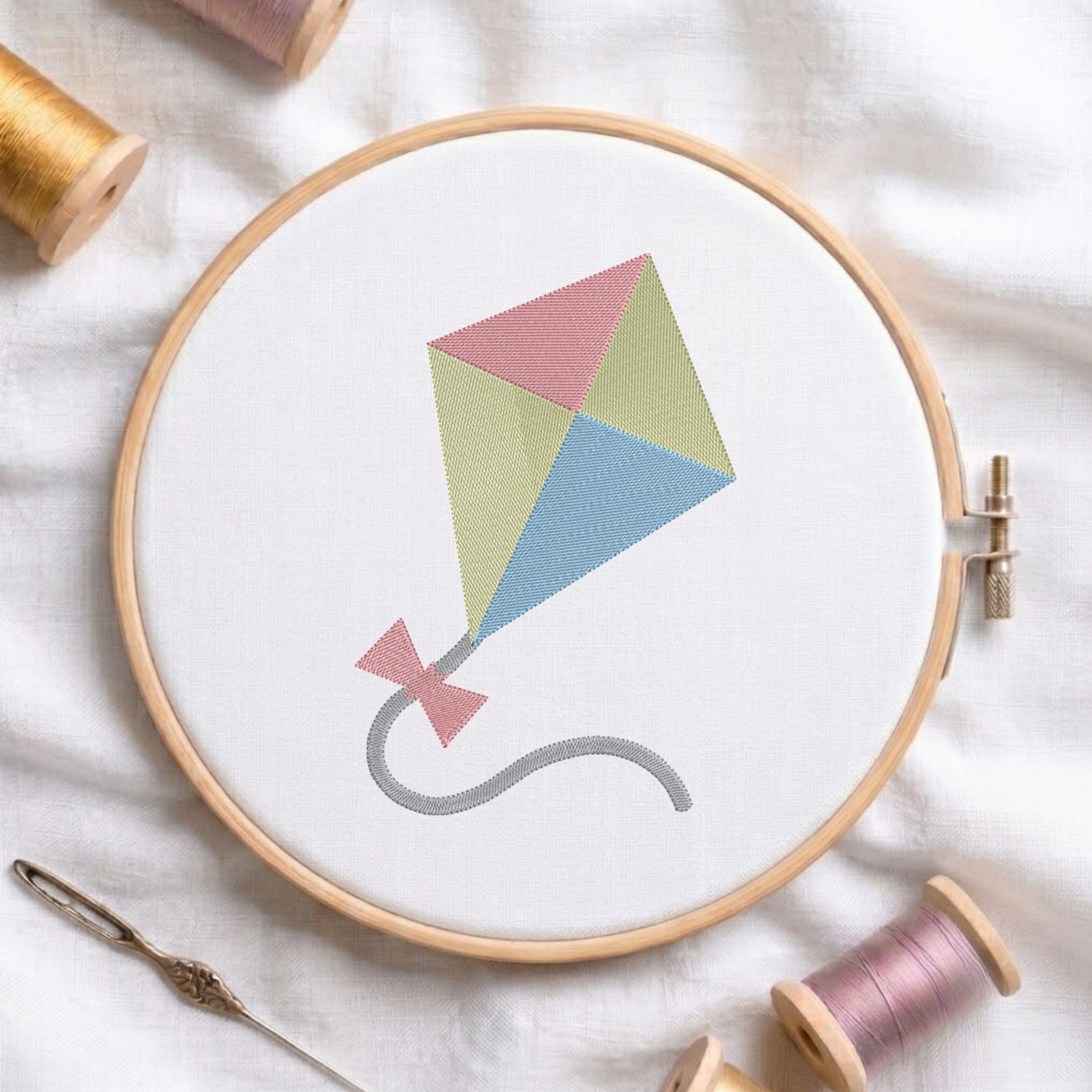 An embroidered colorful kite design with pink, yellow, and blue panels stitched inside a wooden hoop on white fabric with sewing threads.