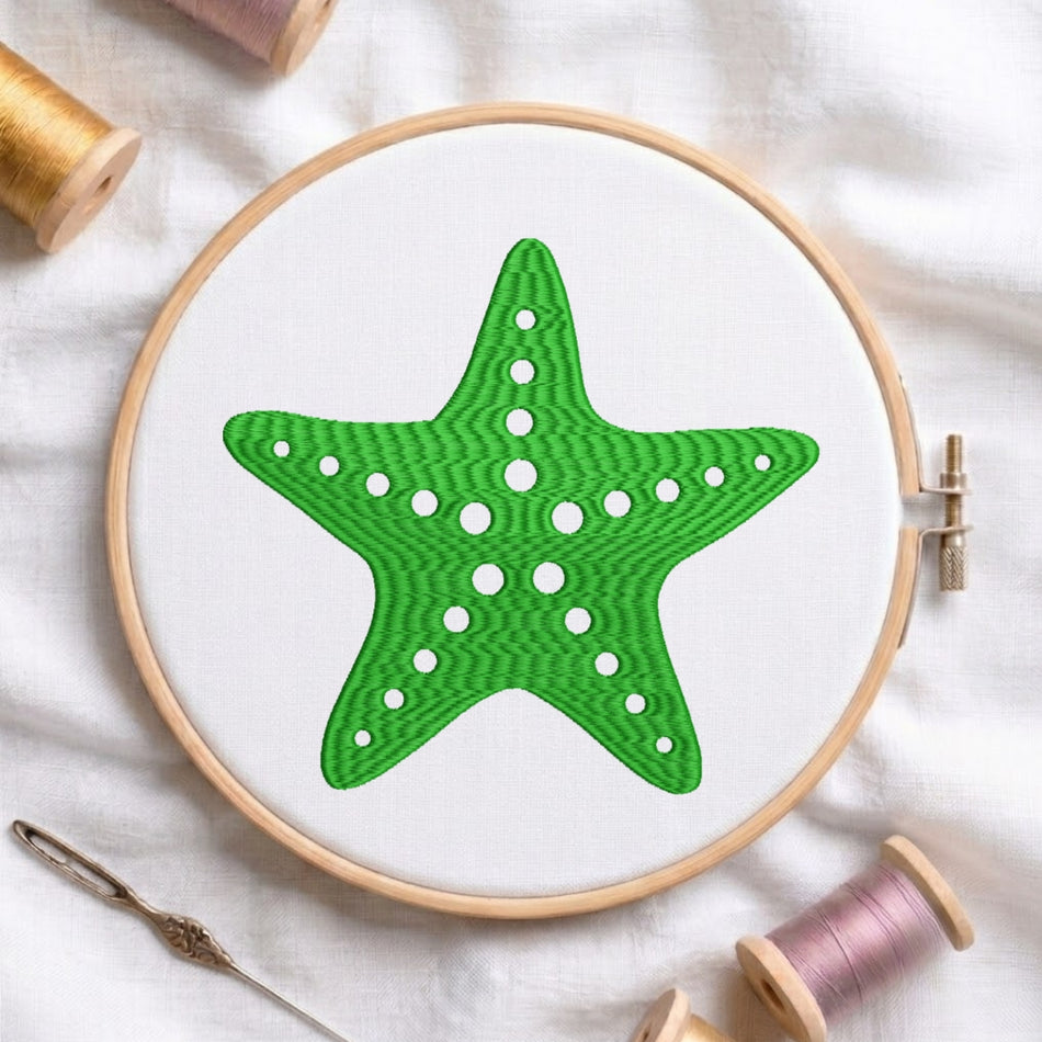 A green starfish design stitched inside a wooden embroidery hoop on white fabric, surrounded by spools of thread and a needle on a white cloth background.