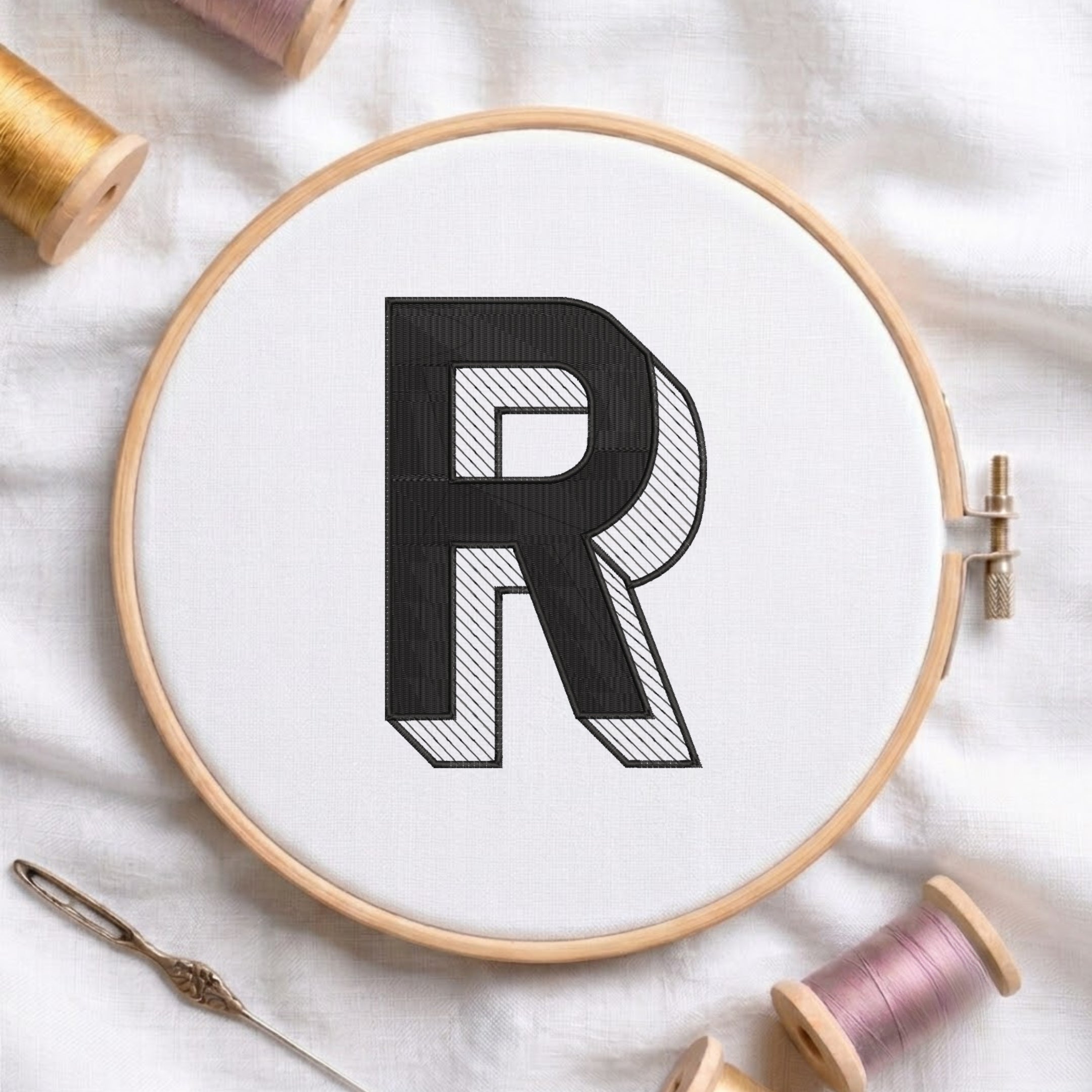 An embroidered 3D block letter "R" with striped shading texture, stitched inside a wooden hoop on white fabric with sewing threads.