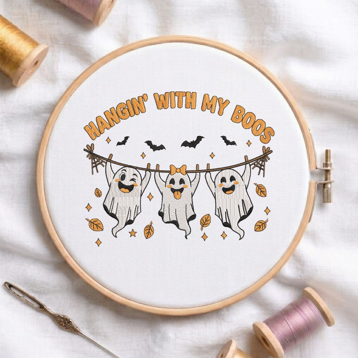 A circular embroidery hoop displays a white fabric with a Halloween-themed cross-stitch design. The design features three smiling ghosts hanging from a clothesline with the text 'HANGIN' WITH MY BOOS' in orange.