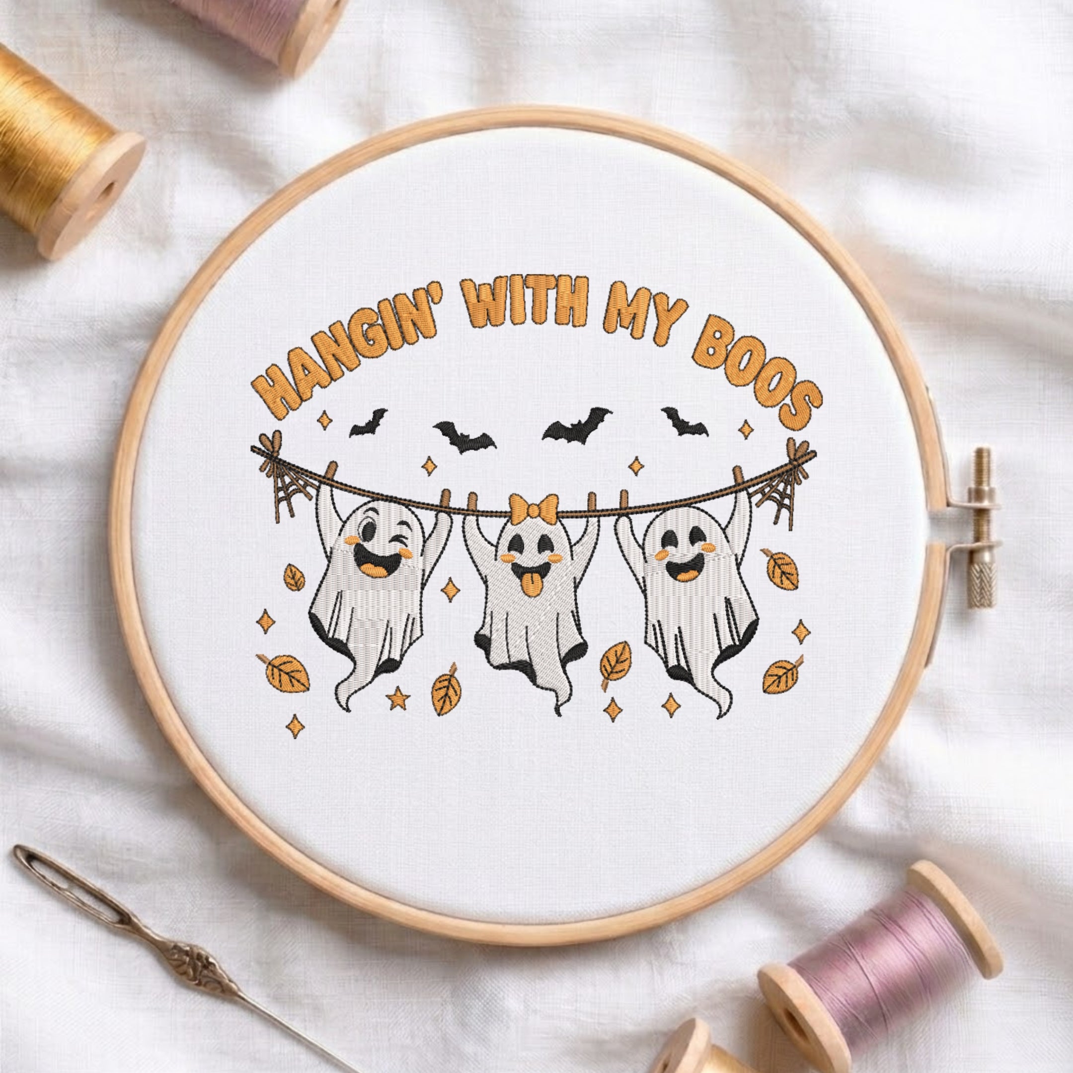 A circular embroidery hoop displays a white fabric with a Halloween-themed cross-stitch design. The design features three smiling ghosts hanging from a clothesline with the text 'HANGIN' WITH MY BOOS' in orange.