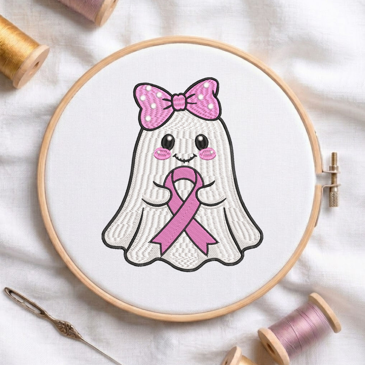 A wooden embroidery hoop holds a white fabric panel with a cute embroidered ghost design wearing a pink polka-dot bow and holding a pink ribbon. The hoop is photographed on a white fabric surface with spools of thread and a needle around it.