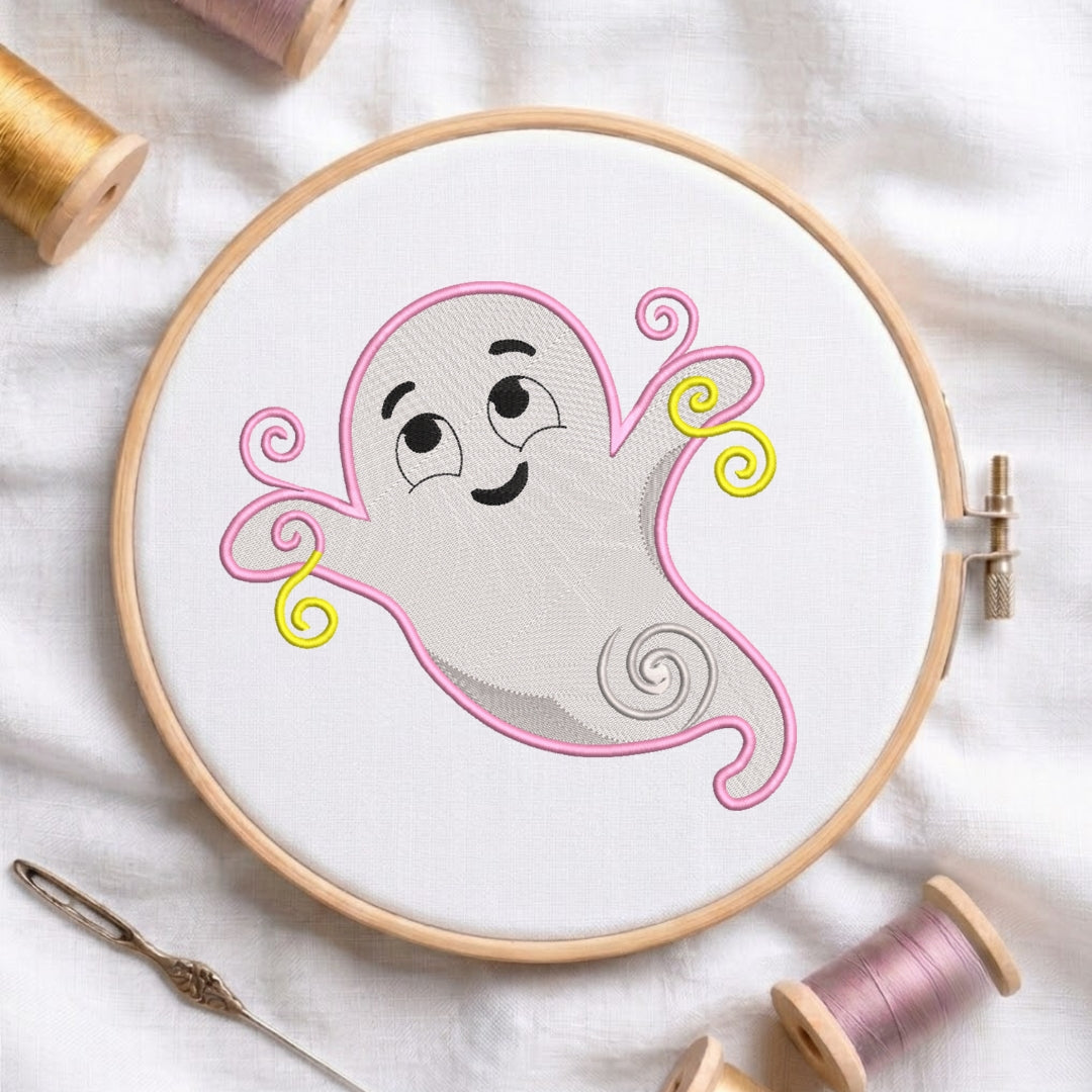 A circular embroidery hoop holding a white fabric panel with a cartoon ghost design outlined in pink and yellow, surrounded by spools of thread and a needle on a white fabric background.