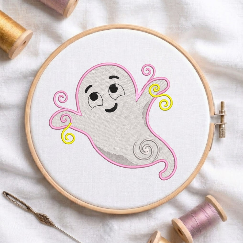 A circular embroidery hoop holding a white fabric panel with a cartoon ghost design outlined in pink and yellow, surrounded by spools of thread and a needle on a white fabric background.