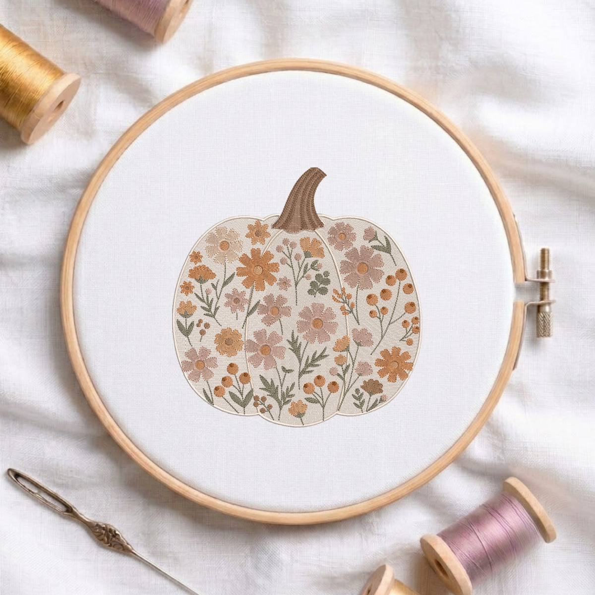 A wooden embroidery hoop holds white fabric with a floral pumpkin design in muted orange, beige, and olive tones. Spools of thread and a needle are arranged around it on a soft white fabric background.