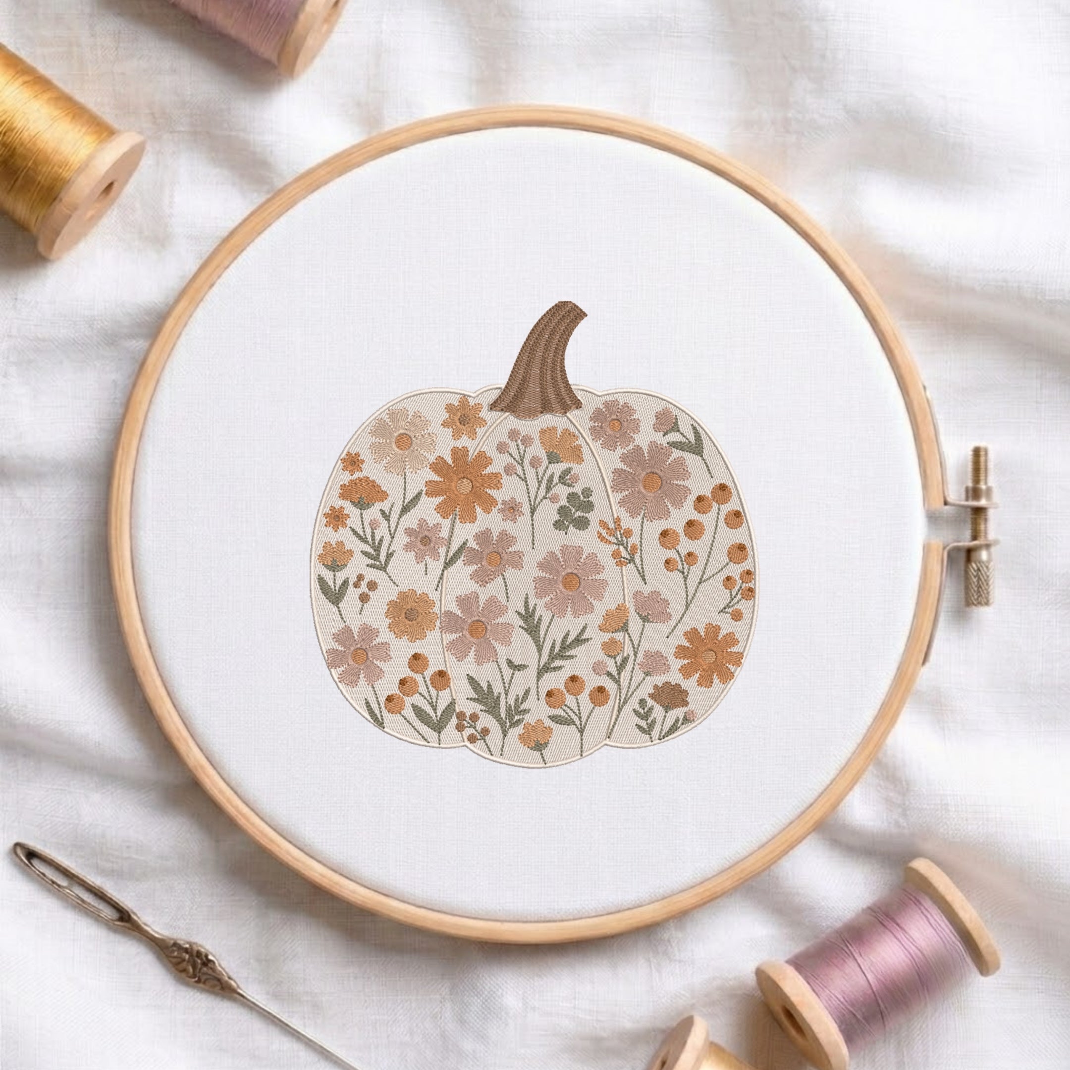 A wooden embroidery hoop holds white fabric with a floral pumpkin design in muted orange, beige, and olive tones. Spools of thread and a needle are arranged around it on a soft white fabric background.