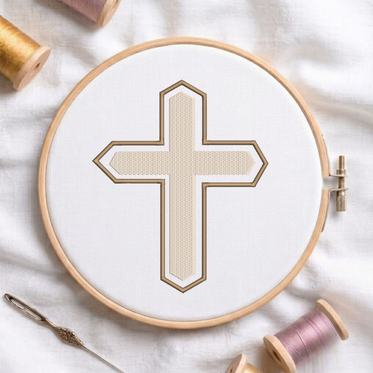 An embroidery hoop on a white cloth displays a finished embroidery design of a textured beige Christian cross with a brown outline, flanked by spools of thread and a needle.