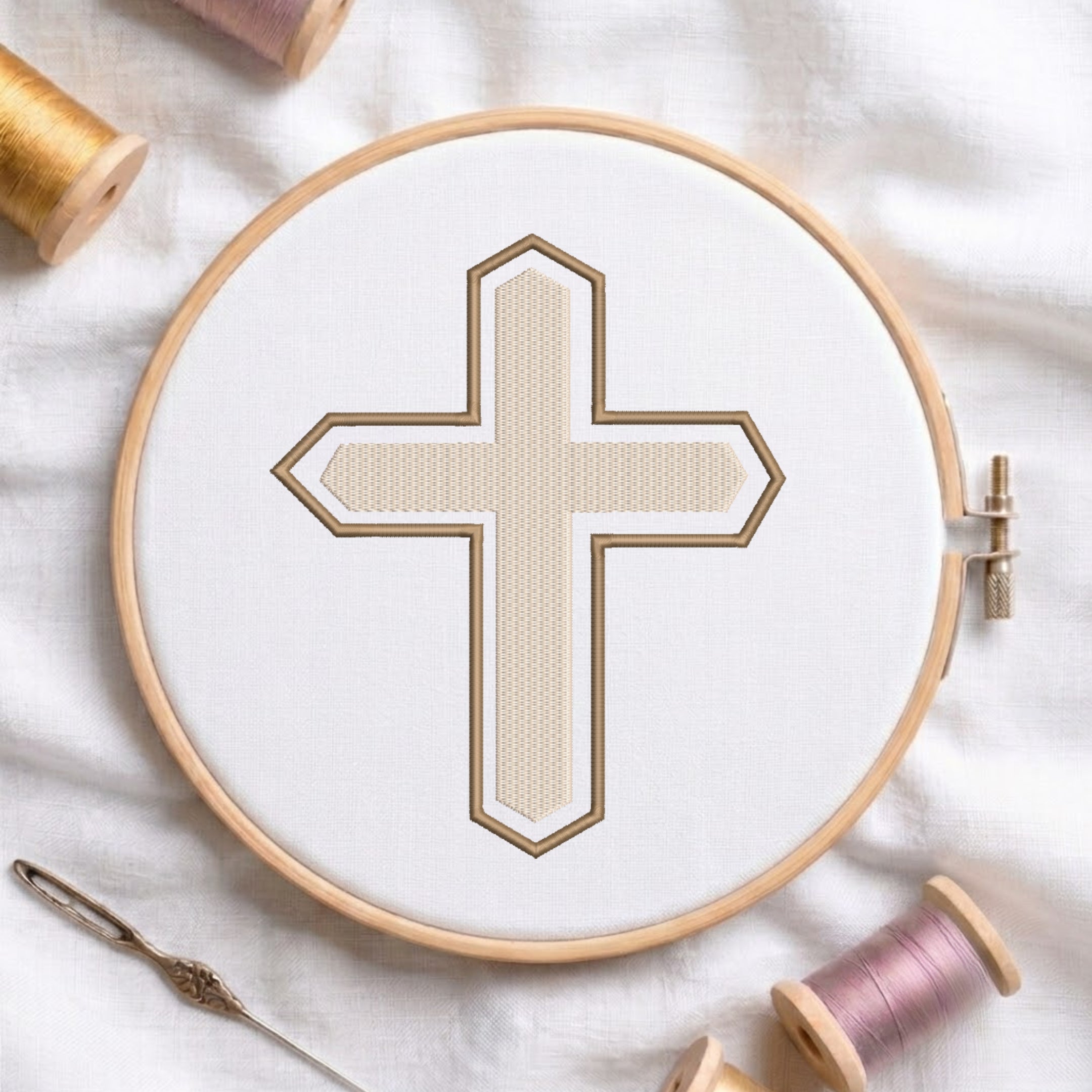 An embroidery hoop on a white cloth displays a finished embroidery design of a textured beige Christian cross with a brown outline, flanked by spools of thread and a needle.