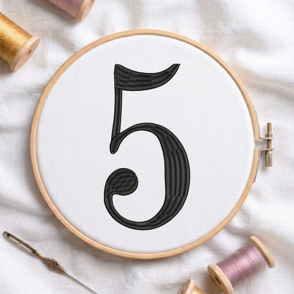 A circular embroidery hoop holding white fabric with a large black stitched number 5 in the center. The hoop is styled on a white fabric surface with spools of thread and a needle around it.