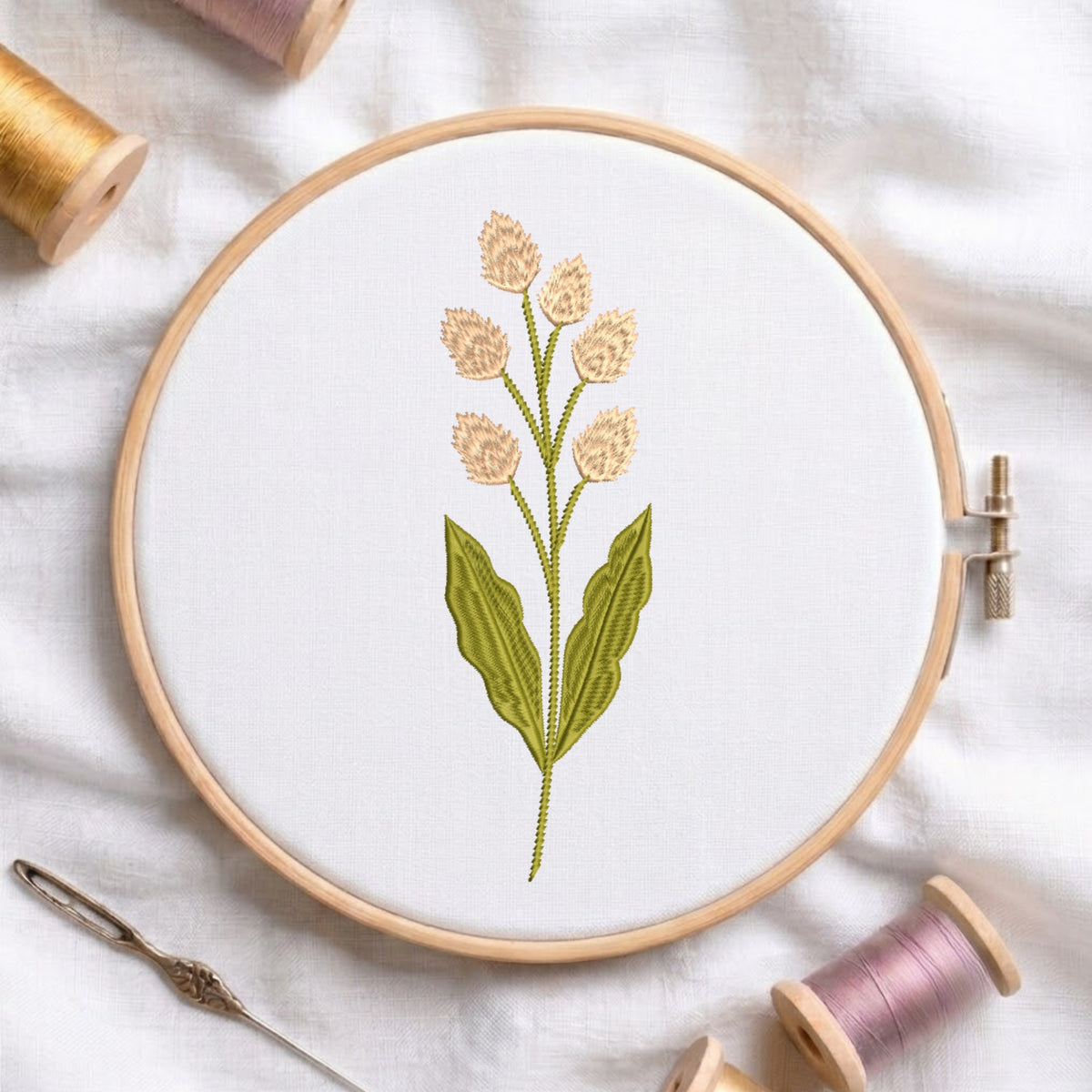 A round embroidery hoop holding white fabric with a stitched floral design featuring beige flowers and green leaves, surrounded by spools of thread and a needle on a white fabric background.