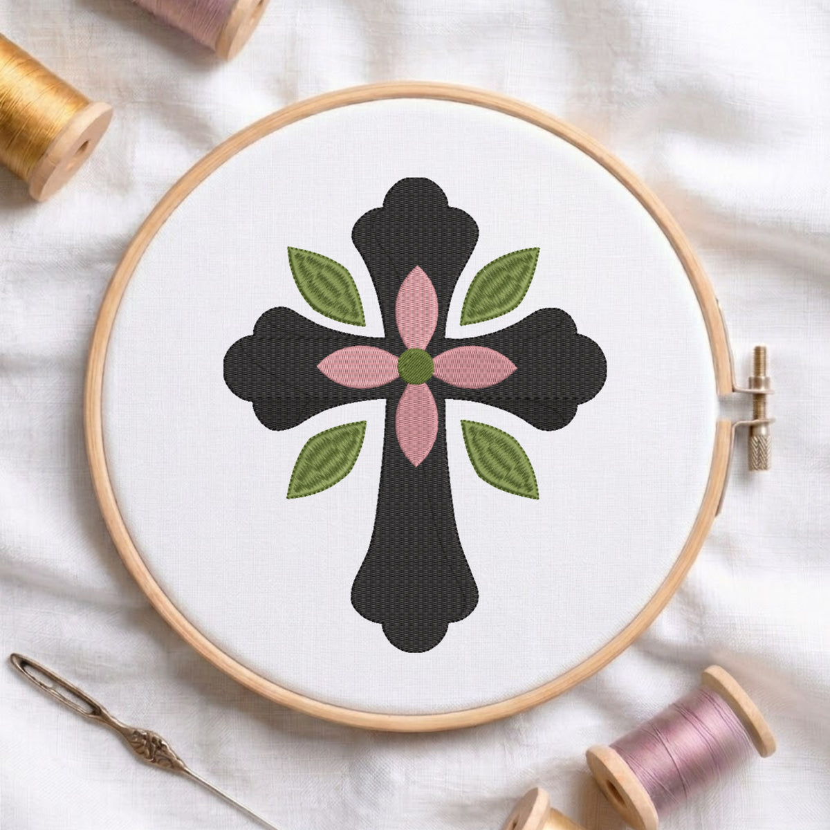 Dark charcoal cross with center pink flower and green leaves embroidered on white fabric inside a wooden hoop.