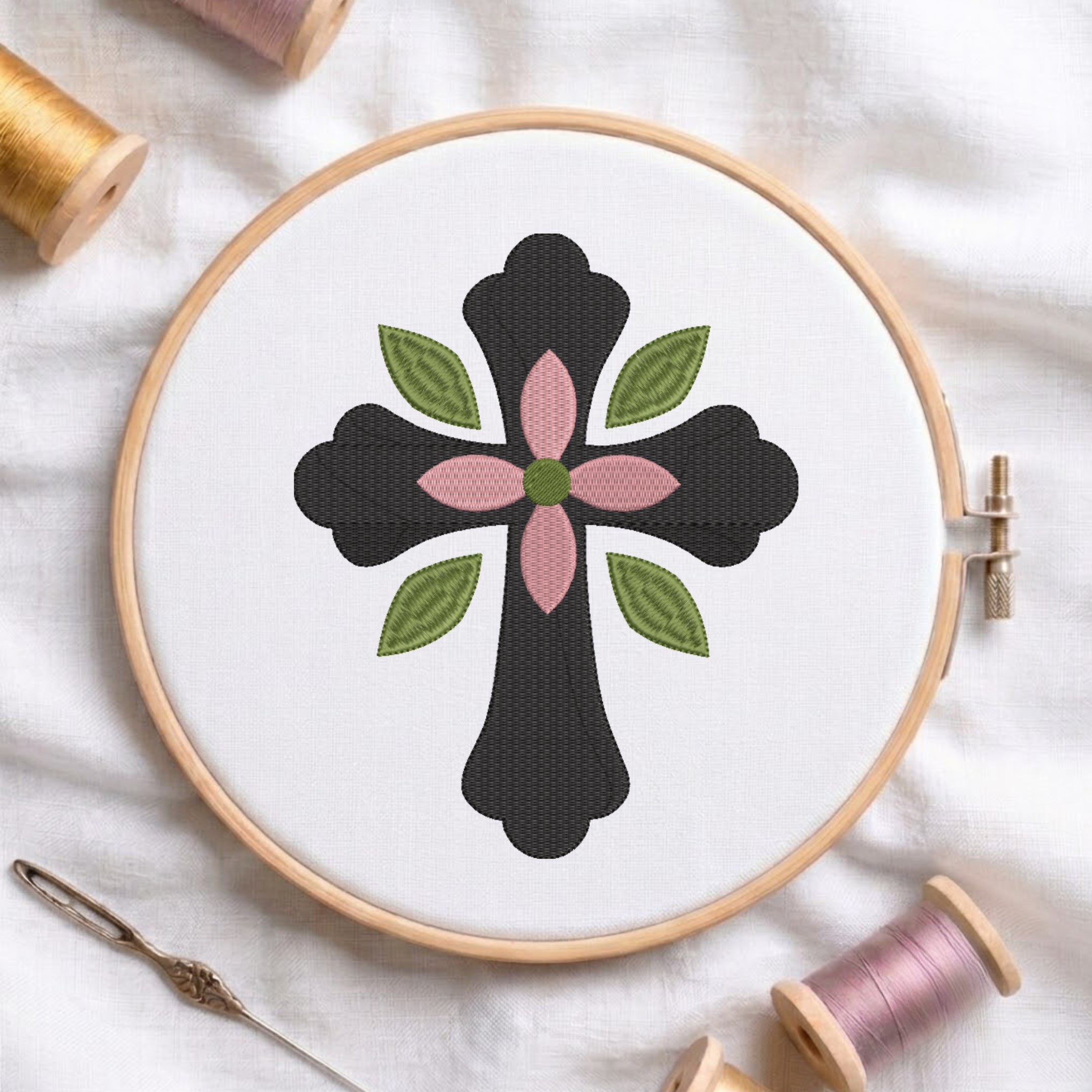 Dark charcoal cross with center pink flower and green leaves embroidered on white fabric inside a wooden hoop.