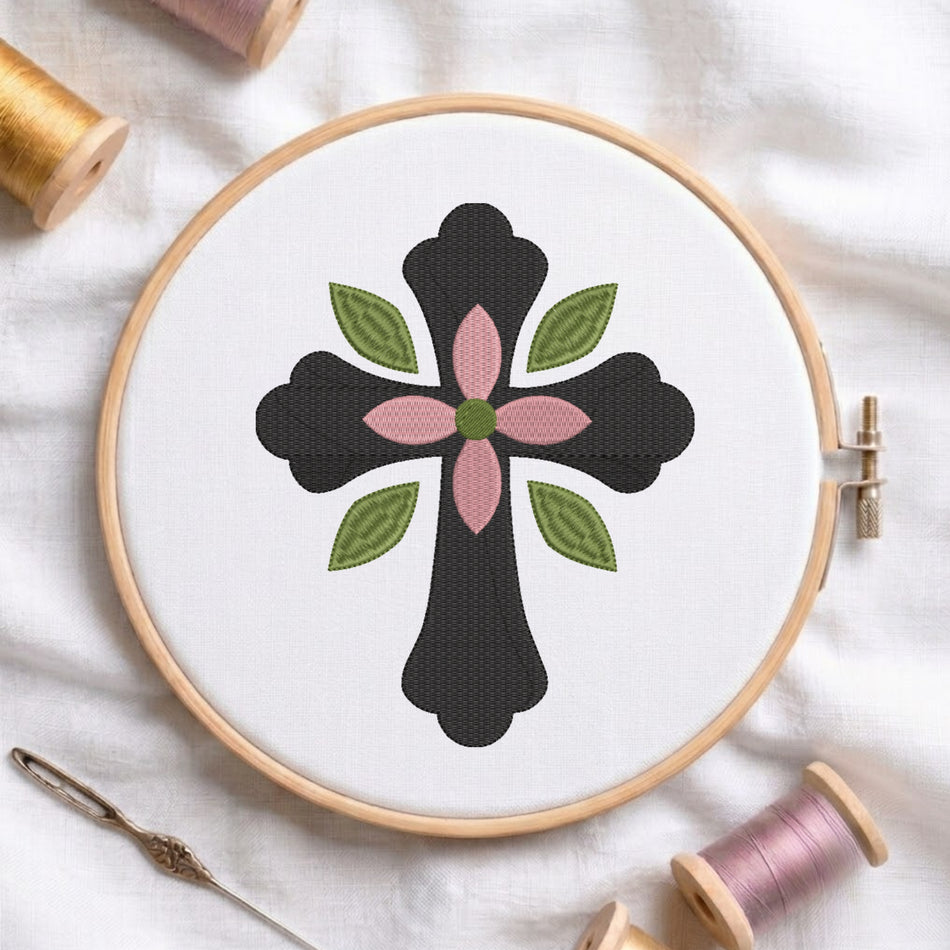 Dark charcoal cross with center pink flower and green leaves embroidered on white fabric inside a wooden hoop.